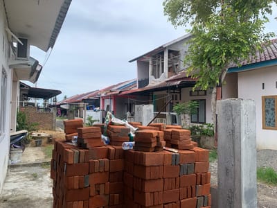 The image, taken in Blangbladeh, Indonesia, shows a residential street with several single-story and partially constructed two-story houses under a cloudy sky. In the foreground, a large stack of red and gray bricks is piled on a concrete path. Litter, including plastic water bottles and white bags, sits on top of the bricks. A weathered, gray concrete pillar stands to the right of the brick pile.
In the background, a row of houses stretches into the distance. Some houses have visible renovations or additions, including exposed wooden beams and scaffolding. A few windows have decorative iron bars. A small tree stands near one of the houses. A motorcycle is parked near a white building in the middle ground. The overall scene depicts a neighborhood undergoing construction and improvement.