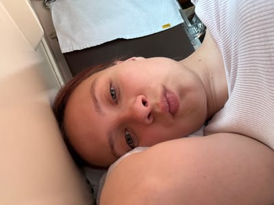 The media file is a selfie taken by a young woman lying down. She is the sole subject and her face is tilted towards the camera. Her eyes are open and looking slightly away from the lens. She appears to be resting on a pillow or soft surface, and is wearing a light-colored, ribbed garment. In the background, a white towel is visible, hanging from a rail or hook. Part of a dark brown surface, possibly a seat or table, is also visible behind the towel. There is a small yellow tag on the towel with some text, which appears to be "PTK 6322". The overall lighting suggests it is daytime, and the scene appears to be indoors, possibly in a mode of transport like a train or bus, given the presence of the towel and hanging rail, and the confined space suggested by the cream-colored surface on the left which could be a wall or seat. The woman's expression is neutral, perhaps a little weary. There is no indication of the weather or a specific time of day beyond it being light.