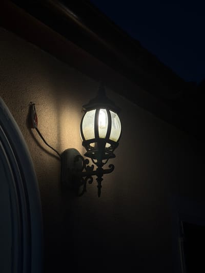 An outdoor, vintage-style wall lantern illuminates a textured wall in Novosilka, Ukraine. The lantern, with a spherical glass shade and decorative black metalwork, casts a warm, yellow light. A red electrical box is mounted on the wall to the left of the lantern, with a black wire connecting it. To the left, a portion of a white, arched architectural feature is visible, possibly a doorway or window frame. Above, dark eaves of a building are silhouetted against a dark blue night sky. The scene is predominantly dark, with the light from the lantern creating a pool of illumination on the wall.