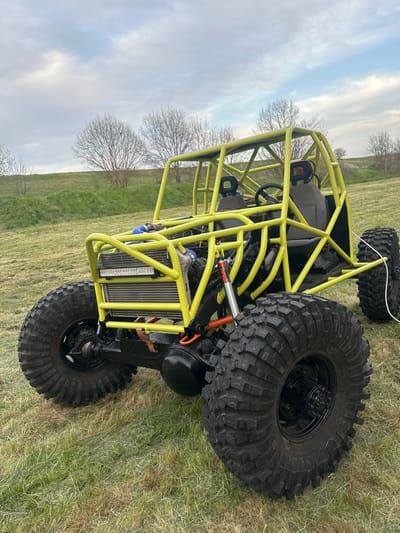 A yellow tubular frame all-terrain vehicle sits on a grassy field in an indeterminate location that is not identifiable as Choba, Bulgaria. The vehicle has oversized black tires, a front-mounted radiator, and two bucket seats. The background includes a horizon line of bare trees against a cloudy sky.