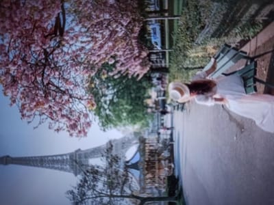 The image depicts a scenic view with the Eiffel Tower in the background, partially obscured by blooming pink trees. In the foreground, a woman in a white dress and a straw hat sits on a bench, her back to the viewer. She is holding something in her hand. The scene appears to be a park or garden setting with a paved walkway. There are other people and a white van in the background, suggesting a public space. The image is taken from a slightly tilted angle.
