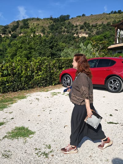 A woman with auburn hair is walking on a gravel driveway in Pozzuoli, Italy, with a red car parked behind her. The woman is wearing a brown top and dark pants, and carrying a phone and a book. The background features a lush green hillside and a building with a balcony. The sky is clear and blue.