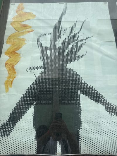 The image shows a poster adhered to a glass surface. The poster features a graphic design dominated by a halftone pattern in black and white. The central image appears to be a stylized depiction of a figure with outstretched limbs. At the bottom of the poster, there are abstract brushstrokes of a golden-yellow color, added on the paper. A human figure with a device or cellphone is reflected in the glass surface, in the upper right side of the image. The reflection overlaps the printed poster design. The location is probably in the city of Arrecife, Spain because some of the printed letters written on the poster are mirrored ("MEDIAZ", "YTUA") suggesting it's a mirrored image and this poster is part of a street ad campaign from a media company, located in the city of Arrecife, Spain.