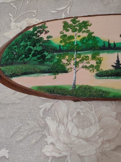 The image displays a framed artwork, possibly a painting or a mosaic, depicting a serene natural landscape. The artwork features a body of water, a tree with green foliage in the foreground, and a distant treeline against a pale yellow and green sky. The frame appears to be made of wood with a natural, unfinished edge. The artwork is positioned against a textured wallpaper with a floral pattern in a light grey or beige color. The focus is on the artwork itself, with the wallpaper serving as a background. The location in the city of Шопино, Russia, is not visually discernible from the provided image.