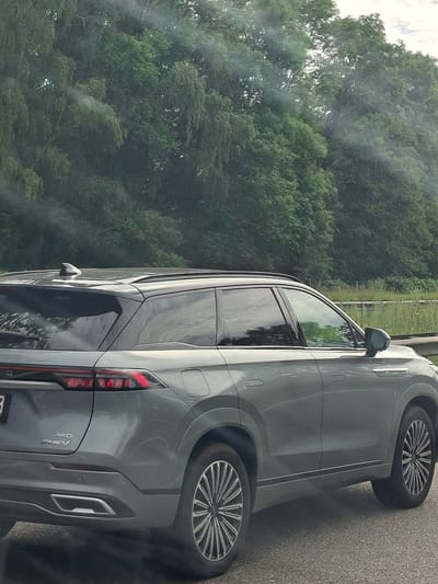 A grey SUV is parked on the side of a road, with trees and a green grassy area in the background. The image is taken from the side of the vehicle, showing its profile. The SUV has tinted windows and distinctive silver wheels. The rear taillight is illuminated red. The sun appears to be out, but the sky is overcast with clouds, suggesting a cloudy day. There is some smoke or haze around the rear of the car, potentially from exhaust or a nearby industrial process. Visible text on the back of the car includes "AWD" and "THEV", indicating it's an all-wheel-drive plug-in hybrid electric vehicle. The setting appears to be Zolder, Belgium. The image is oriented vertically.