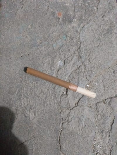 A single, partially smoked cigarette lies on a cracked, grey concrete surface. The cigarette is oriented diagonally, with the filter end pointing towards the right and slightly upwards. The tobacco end is to the left, darker and slightly charred. The filter is white and appears to be made of paper. The concrete surface shows signs of wear and tear, with numerous cracks and some small debris scattered across it. A dark shadow is cast on the left side of the image, possibly from an object or person outside the frame. The scene is outdoors in what appears to be a public space, given the concrete ground.