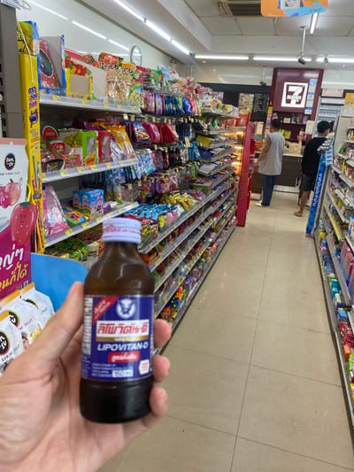 The image shows a hand holding a small brown bottle labeled "LIPOVITAN-D" in Thai, in front of a 7-Eleven store in Ban Ko Pru, Thailand. The shelves are filled with various snacks and candies. Two customers are visible in the background aisle near the 7-Eleven signage. The store has a tiled floor.