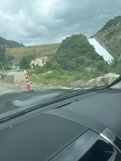 The image, taken from inside a vehicle, shows a view of the Zhinvali Dam in Zhinvali, Georgia. The dam is a prominent feature, with a large, sloping concrete spillway descending from the top of the dam. Water appears to be flowing down the spillway, though the exact volume is unclear. The surrounding landscape consists of steep, green, tree-covered hills and mountains, with a cloudy, overcast sky. In the foreground, part of the vehicle's dashboard and windshield wipers are visible. A red and white striped traffic cone is positioned on the roadside, indicating potential construction or a hazard. A building, possibly a power station or related infrastructure, is visible on the hillside below the dam. The terrain is rocky and appears to be undergoing some form of construction or maintenance.