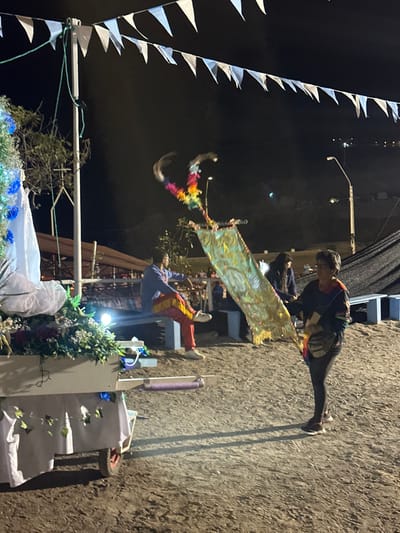 A nighttime scene with two people on opposite sides of a shimmering banner. One person stands on what appears to be a ladder, wearing colorful pants and a blue top. The other person stands above, wearing dark clothing with a colorful design. Pennant banners frame the left side of the image against a dark sky. Below, a decorated structure with flowers and greenery adds to the festive atmosphere.