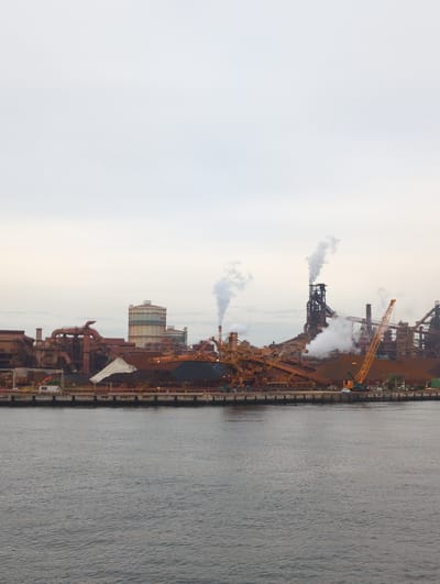 The image depicts a sprawling industrial complex situated along a body of water, likely a port, in Nagoya, Japan. The scene is dominated by large-scale industrial structures, including towering stacks, cranes, and a network of pipes and machinery, all rendered in shades of rust and metal. Several of these structures are actively emitting plumes of white steam or smoke into the overcast sky, indicating ongoing industrial processes.

The time of day appears to be either early morning or late afternoon, given the diffused light and muted colors of the sky. The weather is cloudy, contributing to the somewhat somber atmosphere of the industrial landscape. The water to the right of the complex is calm, with a textured surface reflecting the subdued light.

No people are visible in the image, emphasizing the scale and automation of the industrial operations. The overall impression is one of heavy industry and manufacturing, with the complex stretching along the waterfront. The location is identified as Nagoya, Japan, which is known for its significant industrial and port activities. There is no visible text in the image.