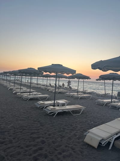 This is a vertically oriented, eye-level photograph of a black sand beach in Rhodes, Greece, during either sunrise or sunset. The scene is dominated by rows of empty white sun loungers and closed umbrellas, arranged neatly on the dark, textured sand. The calm sea stretches out in the background, meeting a pale, gradient sky that shifts from a soft yellow and orange near the horizon to a light blue overhead.
Several figures, appearing as small silhouettes, can be seen near the water's edge, suggesting a few people are enjoying the beach despite the apparent early or late hour. The overall mood of the image is tranquil and serene, with the quiet beach and the soft glow of the sky creating a peaceful atmosphere. The sun loungers are made of a light-colored material, possibly canvas or a similar fabric, and are set on white metal frames. The umbrellas are closed, their fabric a muted gray or off-white. The black sand is uneven, showing natural ripples and indentations. A single visible logo on one of the sun loungers appears to be a circular emblem with some text, but it is too indistinct to be read.