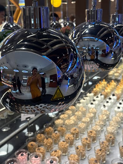 Two silver, spherical objects with cylindrical attachments reflect the surrounding environment. Reflections include people, a restaurant setting, and a warning symbol. A grid of small glass cylinders containing light brown and white objects is visible in the background. The cylinders appear to be arranged in rows and columns, possibly as a display or decoration. The objects have a consistent shape with circles on the front.