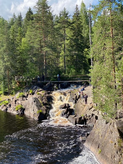 The image depicts a scenic waterfall in Ruskeala, Russia, a town renowned for its marble canyons. A tiered waterfall cascades down dark, rocky cliffs into a dark, rippling body of water in the foreground. Several people are visible in the scene, some standing on the rocks near the waterfall, others on a suspension bridge that spans the river upstream. The bridge is made of wood and rope, with wooden railings. A wooden viewing platform is also visible to the left of the bridge. The surrounding environment is a dense forest of tall pine trees and other deciduous trees under a partly cloudy sky. The water is a murky brown, likely due to sediment from the waterfall, and white foam is visible on its surface. The rocks are rugged and uneven, covered in patches of green moss and grass. The overall impression is one of a natural, rugged landscape enhanced by man-made structures for visitor access.