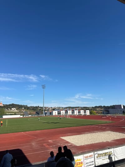 The image shows a view from a stadium in Valpaços, Portugal. The stadium has a running track and a green football field. There are people on the track and field. In the background, there are buildings and trees on a hill. A light pole is also visible. A few spectators can be seen in the foreground. A sign with "JUVENAL PADARIA E PASTELARIA FOLAR TRADICIONA" and phone numbers "933 115296" and "278 996 096" is attached to the stadium. "TAKE-AWAY ENTREGAS AO DOMICILIO" is also on the sign. Another sign says "HOSPITAL MISERICÓRDIA". The sky is blue with some clouds.