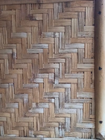 The image depicts a close-up view of a woven bamboo wall panel, likely within a structure located in Kyauk Hsoke, Myanmar. The panel exhibits a diagonal basketweave pattern composed of interwoven bamboo strips varying in color from light tan to medium brown, with some strips showing lighter, weathered tones. A vertical, thicker bamboo structural pole is visible along the right edge, framing the woven panel, with dark circular fasteners intermittently visible on its surface. No people, actions, or interactions are present in the scene. The environment suggests an interior or sheltered space utilizing natural construction materials.