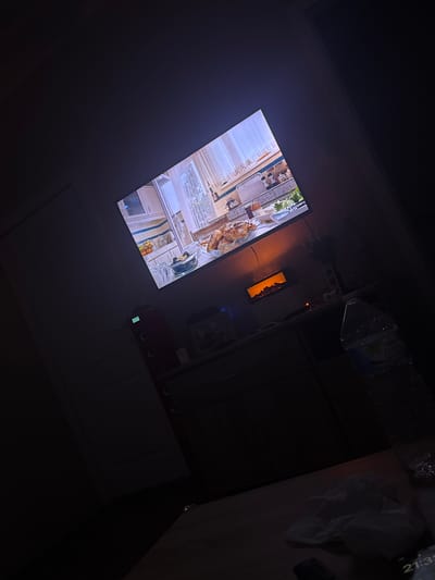 The image depicts an interior scene in Temse, Belgium. A television screen displays a cooking show. Next to the TV, there is a built-in electric fireplace on the wall. A kitchen cabinet is on the right side of the fireplace. Above the cabinets, there are plastic bags. A white plastic bag and a black remote control are on the surface. The room is dimly lit.