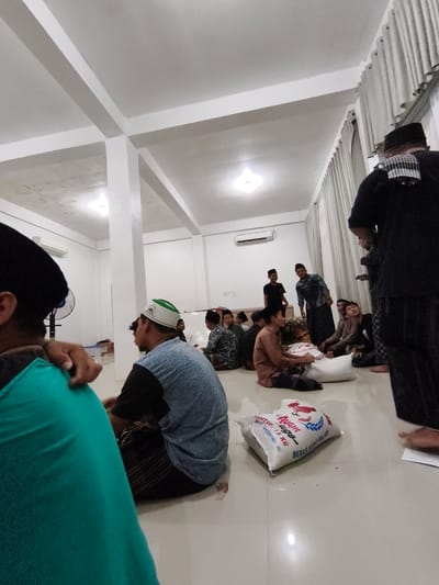 A group of men are gathered indoors, seated on the floor in what appears to be a communal space or prayer hall. The setting is brightly lit, with white walls and a white ceiling featuring prominent beams. Several individuals are wearing traditional Islamic attire, including peci caps and sarongs.
In the foreground, the back of a person wearing a teal garment is visible. To their right, a man in a blue shirt and a white peci is seated. Further into the scene, a group of men are interacting. One man, standing in a patterned shirt and sarong, is gesturing towards a pile of white bags on the floor. Another man, in a brown shirt, is seated and appears to be involved in handling these bags.
On the floor, a white bag is prominently displayed with visible text. The text reads "Ayam Jago" and also "BERAS KETUPAT" and "15 KG". The bags on the floor likely contain rice, suggesting the distribution of aid or food.
The atmosphere appears to be calm and communal. The lighting suggests it could be daytime, though indoor lighting is the primary illumination. The presence of an air conditioning unit on the wall indicates a warm climate, consistent with the location context of Pulodang, Indonesia. The overall scene suggests a community gathering, possibly for a religious event or a charitable distribution.