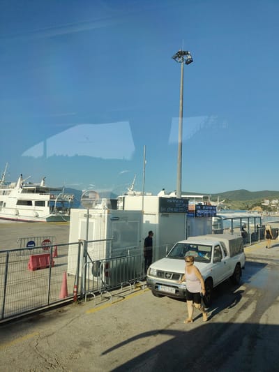 A white pickup truck with a beige canvas canopy is parked on a paved area next to a metal fence. A woman with blonde hair, wearing a pink striped tank top and black shorts, is walking away from the truck. In the background, several ferries are docked at a port. A tall lamppost with multiple lights stands near the truck. The scene appears to be a ferry terminal in Glyfa, Greece, with a clear blue sky and distant hills.