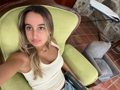 The image captures a young adult female seated in a light green upholstered armchair within an indoor setting in Oliveira de Frades, Portugal. She has light brown hair with blonde highlights and is wearing a light-colored ribbed tank top, with partially visible blue denim bottoms. Her facial expression is characterized by a slight pout, with lips downturned and eyes looking directly at the camera. A dark-covered book, with visible text including "KOL. DECKER" and "Harry Quebert", is held in her lap. To the right of the primary armchair, a second, empty armchair with blue and light-striped upholstery is present. The floor is tiled with reddish-brown square tiles. In the upper right background, an electronic keyboard on a black stand, partially covered by a light-colored cloth, is positioned against a wall constructed from rough stone blocks.