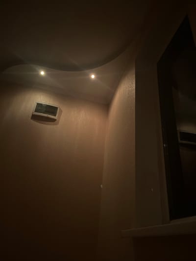The image depicts an interior corner of a room, likely a bedroom or hallway, at night. The lighting is dim and appears to be from recessed ceiling lights and a light source from a window on the right side of the frame. The walls are covered in textured wallpaper, possibly a pale pink or beige color. On the left wall, a square object is mounted, appearing to be a ventilation grille or an old television. The ceiling has a curved architectural feature. A windowsill is visible on the right, with the dark reflection of what might be a window or a dark doorway on the right side. There are no people or visible activities taking place in the scene. The image does not contain any specific landmarks or elements that would allow for its precise location within the city of Pivniceni, Moldova.