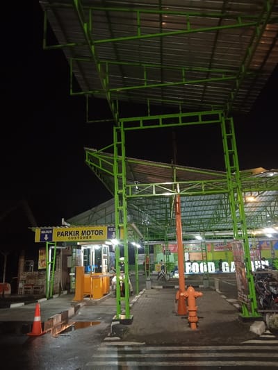 The image depicts a nighttime scene at the entrance of a facility, identified as a "FOOD GARDEN" in Kecamatan Kartasura, Indonesia. The most prominent feature is a large, brightly painted green metal structure that forms an overhead canopy and an entrance archway, supported by an intricate framework of beams. The canopy has a corrugated metal roof.
On the left side of the entrance, a well-lit parking booth with yellow and white accents is visible. A sign above the booth clearly states "PARKIR MOTOR CUSTOMER" (Motorcycle Parking Customer), accompanied by a blue arrow pointing downwards to indicate the entrance. Another sign, partially visible, likely signifies the exit. An orange and white barrier arm is part of the parking system. An orange traffic cone rests on the ground nearby, which shows puddles, suggesting recent rain.
A person, possibly a parking attendant, is visible near the booth in a light-colored uniform. Further inside the facility, additional people and several motorcycles can be vaguely seen.
In the center of the main entrance, a distinct bright orange, pipe-like structure, resembling either a decorative bollard or a fire hydrant, stands out. Beyond the immediate entrance, more green metal canopies and structures extend into the dimly lit background, leading towards a larger area where a prominent sign displays "FOOD GARDEN." Artificial lighting illuminates the structures and the immediate ground, contrasting with the dark night sky. The scene captures the functional operation of a commercial establishment's motorcycle parking entrance after dark.