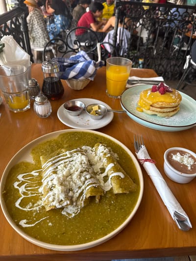 The image shows a table at a restaurant in San Miguel de Allende, Mexico, set with a breakfast spread. The table is wooden and features various items: a plate of enchiladas verdes topped with cream and cheese, a plate of pancakes with raspberries, a side of refried beans topped with cheese, a glass of orange juice, a small dish with butter and jam, and condiments like syrup, salt, and pepper. Cutlery wrapped in a napkin is also present. The setting appears to be outdoors, as parts of people and a wrought iron barrier can be seen in the background. People are seated at a nearby table.