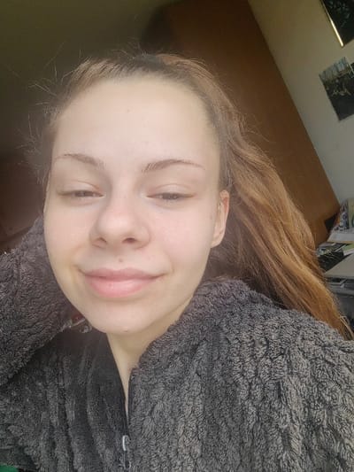 The image depicts a young Caucasian female, approximately late teens to early twenties, located indoors in Dolna Kremena, Bulgaria. She is positioned centrally in a close-up, head-and-shoulders shot, looking directly at the viewer with a slight, closed-mouth smile and partially closed eyes. Her hair is light brown or dark blonde, pulled back from her face with some loose strands visible. She wears a dark grey, textured, possibly fleece, zip-up garment with the zipper partially visible. Her left hand is raised to the left side of her head, partially obscured by her hair. The background on the right consists of a light brown panel or furniture, with two framed pictures or posters mounted on a wall above it, and an indistinct stack of objects below. The background on the left is darker and less defined, suggesting a shadowed area or another piece of furniture.