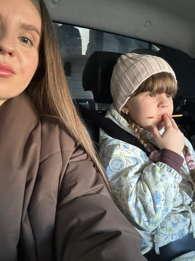 This image depicts two individuals inside a passenger vehicle in Konakovo, Russia. On the left, a woman's partial face and upper body are visible, showing her left eye, nose, and part of her mouth with light pink lipstick. She has long, straight light brown hair and is wearing a dark brown, possibly quilted, jacket. Her gaze is directed towards the camera.

To her right, a young child is seated, secured by a black seatbelt across the chest. The child wears a light pink ribbed knit beanie and a light-colored winter jacket featuring a floral pattern of blue and green leaves/flowers on an off-white background. A circular white patch with the word "FUTURINO" and a heart symbol is affixed to the jacket's left chest. The child has shoulder-length light brown hair styled in two braids, each secured with a pink scrunchie or tie. The child is holding a thin, light-colored snack stick with their right hand, placing it into their mouth. Both individuals are positioned in what appears to be the back seat, with dark headrests visible behind them. A blurry exterior view is visible through the car window in the background.