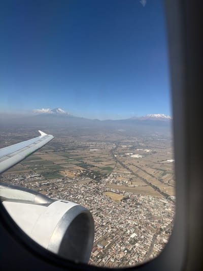An aerial photograph, taken from an airplane passenger window, depicts the landscape above the city of Santa Ana Xalmimilulco, Mexico, in the state of Puebla. The image frames a portion of a white aircraft wing with green detailing on its leading edge in the upper left, and a segment of a white and silver turbofan engine nacelle in the lower left, both partially obscured by the dark, curved window casing. Below the aircraft, the ground reveals a dense urban area, characterized by numerous light-colored, closely packed buildings, which corresponds to Santa Ana Xalmimilulco and its adjacent settlements. This urban development is interspersed with and transitions into expansive agricultural fields, organized into distinct rectangular and irregular plots of varying hues, indicating different cultivation or fallow states. In the distant background, under a clear blue sky with some atmospheric haze, two prominent snow-capped volcanoes are visible: Iztaccíhuatl to the left, partially covered by clouds near its summit, and Popocatépetl to the right, displaying a more defined snow cap. The entire scene is brightly illuminated by daylight. No individuals, specific actions, or direct interactions are visible.