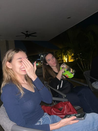 Two women are sitting outside at night. One woman holds her hand over her face and smiles broadly. She wears a blue top and jeans. The second woman sits behind her, holding a bag of nuts and looking at the camera. She wears a black top. A phone and a red bag sit on the chair beside her. A building and palm trees are visible in the background.