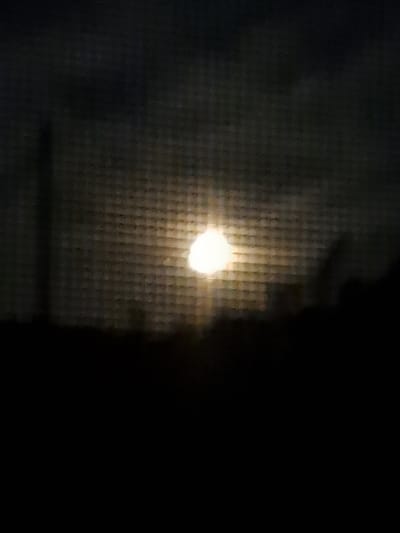 The image captures a dimly lit scene, possibly at dusk or night, through a window screen, as indicated by the grid pattern overlaying the view. A bright, circular light source, likely the moon or a distant streetlamp, is visible in the upper center of the frame, emitting a soft glow. Silhouetted against this light are dark, amorphous shapes suggesting trees or buildings forming a low horizon. A faint, elongated dark vertical shape is also present on the left side of the image, possibly a pole or a thin structure. The overall atmosphere is muted and somewhat abstract due to the low light and the screen's texture. The location in Chernetsk, Russia, is not discernible from the visual information provided in the image.