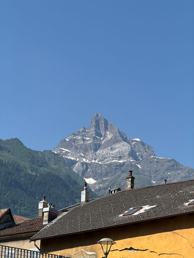 The image shows a view of a mountain with snow-capped peaks against a clear blue sky. In the foreground, there are rooftops of buildings with chimneys. The mountain is rocky and appears to be a significant natural landmark. The overall scene suggests a mountainous region, possibly near a town or village. The image is captured during daylight.