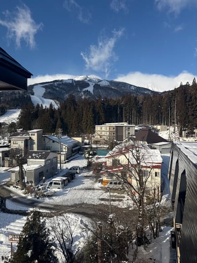 A winter scene shows a snow-covered mountain with ski runs nestled alongside a forested area. Buildings are scattered in a small town below, with snow-lined roads and parked vehicles. The sky is blue, dotted with clouds. The image appears rotated.