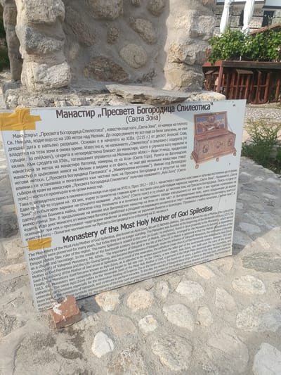 The image shows an informational sign with text in Bulgarian and English, placed on a cobblestone ground in front of a stone structure. The sign's title reads "Monastery 'Presveta Bogoroditsa Spileotisa'" (Monastery of the Most Holy Mother of God Spileotisa). The English text describes the monastery's location on a plateau 100 meters above Melnik, Bulgaria, and its historical significance. The sign is white with black text and an image of an ornate chest. The foreground is dominated by the cobblestone ground and the sign, while the background shows a stone wall and some greenery. There are no people visible in the image. The scene appears to be an outdoor historical site.