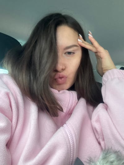 A single female subject with long, dark hair, partially covering the left side of her face, is depicted in what appears to be the interior of a vehicle. Her lips are pursed forward. She is wearing a light pink, textured fleece garment with a visible zipper detail around the collar area. Her right hand is raised, with fingers extended towards her temple; the fingernails are painted white with a pointed shape. The background consists of dark and light gray tones, consistent with a vehicle's headliner and interior trim. No identifying markers for the specific city of Рассудово, Russia, or any other precise geographic location are present within the image.