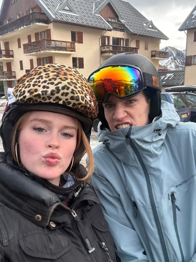 Two individuals, a female and a male, are depicted in an outdoor setting in Saint-Sorlin-d'Arves, France, with alpine resort buildings and snow-capped mountains in the background under an overcast sky. The female, positioned on the left, has reddish-blonde hair and wears a black ski helmet covered with a leopard print fabric. She wears gold hoop earrings, a black winter jacket, and is making a pouting expression towards the camera. The male, positioned to the right, wears a black ski helmet with multi-colored reflective goggles. He is dressed in a light blue winter jacket featuring "PROTEST PURE GREEN" branding and is making a grimacing face with his teeth bared. The background consists of several multi-story buildings with light brown facades, dark pitched roofs, and wooden balconies and window shutters. Snow is visible on the building roofs and on distant mountain peaks. A dark vehicle with a roof box is partially visible behind the male subject on the right.