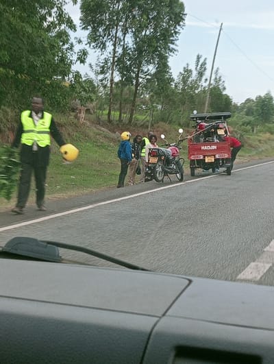 The image depicts a roadside scene in Lwandeti ward, Kenya. The foreground shows the dashboard and windshield wiper of a vehicle from which the photograph was taken. A two-lane asphalt road with a solid white shoulder line extends into the midground.
On the left, a male subject walks on the dirt shoulder, moving towards the viewer. He wears a dark suit or uniform with a bright yellow-green reflective safety vest and holds a yellow helmet in his left hand and green foliage in his right.
Further down the dirt shoulder, a group of four individuals stands near a parked red motorcycle (KMD 8C2X). One person wears a blue top and a yellow helmet; another wears a yellow-green reflective safety vest over a dark top; a third is partially obscured by a yellow-green vest; and a fourth, female, smiles towards the camera.
Adjacent to this group, a red three-wheeled cargo motorcycle (Haojin, KWD 270L) is parked partly on the paved shoulder. Its cargo bed contains at least two motorcycles. A male subject in a red long-sleeved shirt is bent over at the rear of this vehicle.
The roadside is characterized by a dirt and grass shoulder, beyond which tall, slender trees with green foliage line the area. A utility pole leans slightly in the background near the three-wheeled vehicle. The sky appears overcast.