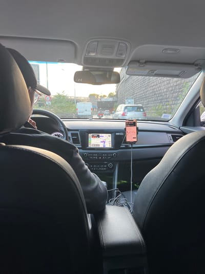 The image depicts the interior of a vehicle viewed from the rear passenger seat, looking forward through the windshield onto a roadway in Le Kremlin-Bicêtre, France. A male driver, visible from behind, wears a dark cap and glasses, with his left hand near his chin. The vehicle's interior features black leather seats, a dashboard with a central infotainment screen displaying a navigation map indicating "5 min / 0.8 km" remaining, and a smartphone mounted to the right of the screen, also showing a navigation or ride-sharing application. Tangled charging cables rest on the black center console between the front seats. A rearview mirror with an "Under Armour" sticker is visible, and the sun visors are stowed.

Outside the vehicle, the scene shows congested or slow-moving traffic under direct sunlight. Vehicles directly ahead include a white delivery van, a silver sedan (likely a Citroën C4 Picasso), and a scooter. To the right of the road, a tall, textured retaining wall constructed of stone or concrete blocks runs parallel to the roadway, featuring a white informational sign. To the left, greenery and a circular "50" speed limit sign are visible. In the distance, an underpass or tunnel entrance is prominent, framed by the surrounding infrastructure. The overall environment suggests a major urban artery or motorway access road, consistent with the A6B (Autoroute du Soleil) or its access ramps near the Boulevard Périphérique and Porte d'Italie area within Le Kremlin-Bicêtre, characterized by its extensive tunnel systems and retaining walls.