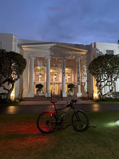 A black Specialized bicycle is parked on a lawn in front of a white neoclassical building at dusk. The building has a grand entrance with large columns, arched windows, and hanging lanterns. The bicycle has a red taillight illuminated, casting a red glow on the grass. A white light from another source illuminates a patch of the lawn to the right of the bicycle. The sky is a deep blue with some clouds. The scene appears to be in Bal Harbour Village, United States of America, likely a hotel or similar establishment given the architecture. There are no people visible in the image.