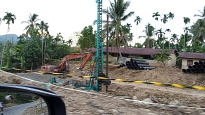 In the city of Buketmulia, Indonesia, a construction site is depicted, featuring an orange excavator positioned to the left, and a large, teal drilling rig to its right. The ground is uneven and covered in dirt and rocks, indicative of ongoing excavation work. Stacked cylindrical black pipes are visible on the right side of the frame, and more are partially visible on a mound of dirt to the left. In the background, several buildings with dark red roofs are present, interspersed with lush green foliage and tall palm trees, characteristic of a tropical environment. A yellow and black striped barrier runs horizontally across the middle of the image, separating the foreground from the construction area. The sky is overcast.