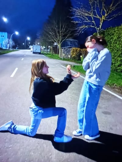 The image depicts a nighttime scene on a paved road in Mirbozor, Uzbekistan. Two young individuals are present. On the left, a person with long, light brown hair, dressed in a dark long-sleeved top, light blue jeans, and white sneakers, is kneeling on their right knee. Their hands are open and raised, palms upwards, facing the second individual. On the right, a second person with dark hair, wearing a light blue hooded sweatshirt, wide-legged light blue jeans, and white sneakers with blue accents, stands facing the kneeling individual. This person has their right hand covering their mouth and nose. The background features a paved road with white dashed lines. Multiple streetlights illuminate the area. To the left, a light-colored building is visible. Further down the road, a white van is present. To the right, a tall green hedge lines the road, behind which are leafless trees and a faint structure. The sky is dark.