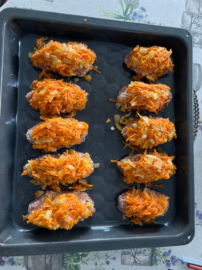 A top-down shot shows a black baking tray filled with ten oblong-shaped portions of meat topped with shredded carrots and diced onions. The tray sits on a table with a patterned tablecloth. The scene is in Лесной Пейзаж Кп (шарапово, Russia.