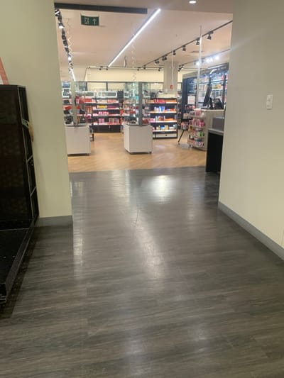 The scene depicts the entrance to an unidentifiable retail establishment, presumed to be located in Lauro de Freitas, Brazil. The foreground features a reflective dark grey wood-grain patterned floor, transitioning to a lighter honey-toned wood-look floor at the store threshold. To the immediate left, a black, open-shelf metal display unit is partially visible and appears largely empty. To the right, a light beige wall with a grey baseboard contains a white wall switch panel.
The store interior is brightly illuminated by linear LED fixtures and multiple track lights mounted on a white ceiling with exposed infrastructure. Two white, square pedestals with dark tops are centrally positioned at the entrance, appearing to function as display units or part of a security gate system. Beyond these, the store contains multiple shelving units along the walls and within aisles, stocked with various cosmetic and personal care products, predominantly in colorful packaging. A checkout counter or service desk is visible on the far right, equipped with a computer monitor. A red fire extinguisher is mounted on a pillar in the right mid-background. A green "EXIT" sign with a right-pointing arrow is affixed to an overhead beam. No individuals are present, and no actions or interactions are observed.