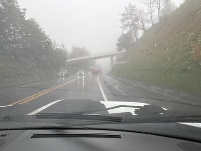 The image captures a rainy, foggy scene from inside a vehicle. Raindrops dot the windshield, obscuring the view of a road with multiple vehicles, including cars and a truck. The road markings are visible despite the weather. A bridge crosses the road further ahead, shrouded in fog. Dense trees and hillside vegetation line the sides of the road, adding to the atmospheric perspective of the inclement weather. The car's dashboard and windshield wiper are visible in the foreground.
