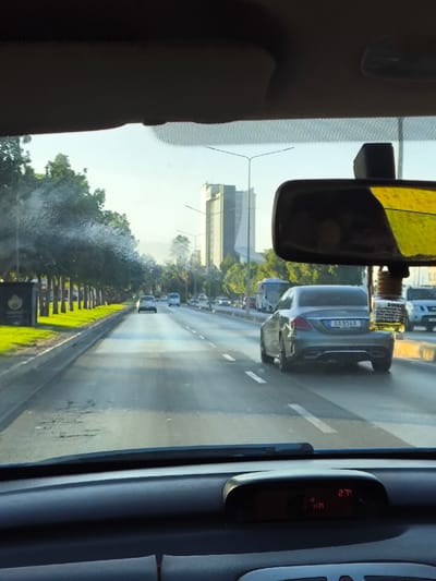 The image depicts a forward view from inside a moving vehicle on a multi-lane road in Gonyeli, Northern Cyprus / Cyprus, under bright daylight. The foreground reveals the vehicle's dark dashboard, with a digital display showing "27° KM". A rearview mirror hangs from the top right of the windshield, reflecting a yellowish tint, and has an air freshener suspended from it, labeled "DİKKAT" (Turkish for "CAUTION") along with smaller Turkish text. The windshield itself exhibits visible streaks and residue, particularly on the left side.

Ahead, a grey Mercedes-Benz sedan, bearing Northern Cypriot license plate "AA 814 A", occupies the right lane. The road surface shows areas of wear and minor cracking. To the left, a landscaped median strip features a row of uniformly spaced trees and green grass, with a dark rectangular sign partially visible. Further down the road, additional vehicles, including cars, a white van, and a bus, indicate moderate traffic flow. Streetlights line the road on both sides. In the background, under a clear sky, a prominent, modern tall building with a distinctive architectural profile stands out against the horizon, characteristic of the city's skyline.