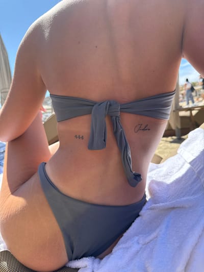 An adult female is depicted from the back, seated in an outdoor, sunny environment in Ornos, Greece. She is wearing a two-piece, solid grey/slate-colored swimsuit. The bandeau-style top is tied in a knot at the center of her mid-back. A small, dark mole is present on her upper back, slightly right of the spine. Two tattoos are visible: the numerals "444" are tattooed in a simple font on her left lower back, and the word "J'adore" in cursive script is tattooed on her right rib cage/side, above the waistline. Faint stretch marks are visible on the upper left gluteal area. She is seated on what appears to be a lounge chair with a light-colored, woven surface. A white terry cloth towel is draped beside her on the right. In the blurred background, a clear blue sky is visible above a beach or resort setting, including a partial view of a light-colored, possibly striped, beach umbrella on the left and a raised wooden structure with blurred figures of other individuals on the right.