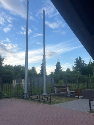 The image depicts an outdoor scene in Dubovaya Roshcha, Russia, likely a park or recreational area, during daytime. Dominating the sky are scattered white clouds against a blue backdrop. Two tall, slender metallic poles are positioned prominently in the foreground, reaching upwards towards the sky. To the right of these poles, a wooden bench with concrete supports is visible, accompanied by a wooden trash receptacle. Beyond the bench, a green metal swing set is present, suggesting a children's play area. A black metal fence encloses the area, separating it from a strip of green grass and a row of trees that form the background. In the far background, a light-colored, rectangular structure with yellow markings, possibly an electrical substation or utility box, can be seen behind the fence. The foreground is paved with red brick tiles, with some grass growing between them. A dark, angled structure, possibly a roof overhang or awning, occupies the upper right portion of the frame.