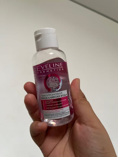 A hand holds a clear plastic bottle of Eveline Hyaluronic Micellar Water. The bottle has a white flip-top cap and a pink label with white text. The label features the brand name "EVELINE COSMETICS" and indicates that the product provides "24H Hydration" with "AquaXyl™ Hydraconcept 3D™" technology. It also states that the micellar water "removes waterproof makeup," "deeply cleanses," and "reduces irritation." The text also mentions it is suitable for "dry, sensitive, and irritated skin" with a "hypoallergenic formula" and "0% alcohol." The bottle is partially obscured by the fingers holding it. The background is a plain white surface. The image does not provide any information about the location in the city of Байсерке, Kazakhstan.