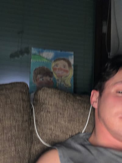 The image presents an indoor scene featuring a partial view of an individual and an artwork. A male individual is visible from the upper right quadrant of the frame, showing his right ear, dark hair, and the shoulder area of a grey shirt. A white earbud cable is draped from his right ear, over his shoulder, and descends into the cushions of a textured, brown upholstered couch on which he appears to be seated. The couch occupies the lower and central-left portions of the frame, with visible backrest and seat cushions. Directly behind the couch and centrally positioned on a dark greenish-grey wall is a colorful, unframed drawing. The drawing depicts two stylized, large-headed cartoon figures: a male with brown hair on the left and a female with blonde hair on the right, both featuring wide smiles and rosy cheeks. The background of the drawing includes green hills, a blue sky with white clouds, and a distant white structure resembling a steeple or tower on the left hill. Above the drawing, a blurred light fixture with three distinct dark, spherical elements is mounted. The overall environment is dimly lit. The specific location in Lengdorf, Austria, cannot be determined from the visual content of the image.