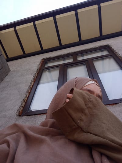 In the city of Suzak, Kyrgyzstan, a low-angle shot captures a person wearing a hijab and a brown abaya. The individual is partially obscured by their clothing, with only their eyes and forehead visible. They appear to be looking upwards, away from the camera. Behind the person, a textured concrete wall is visible, featuring a large window with dark brown framing. The window is divided into multiple panes, with a white screen visible behind the glass. Above the window, a section of a building's overhang is present, with light-colored panels supported by dark beams. The sky is visible in the upper portion of the image, appearing overcast and pale. The overall scene suggests an outdoor setting against a building facade.